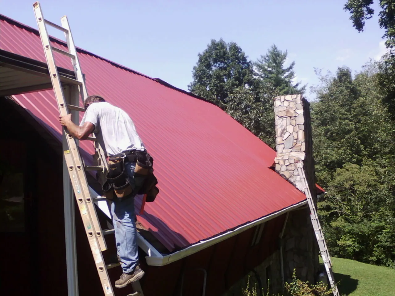 Roofer on a ladder beside a red metal roof and stone chimney — Soffit & Fascia Repair by Roofing Brothers in Chula Vista